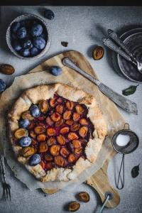 A plum pie on a wooden board on concrete backdrop with utensils
